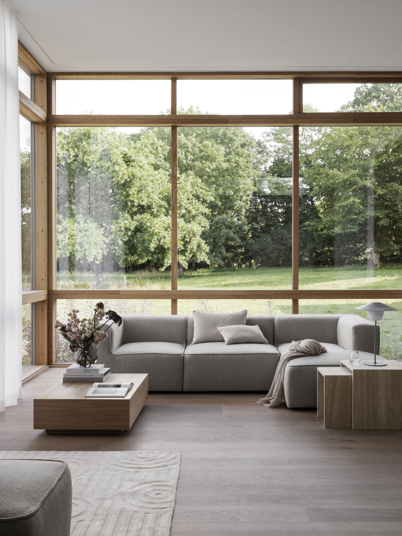 Moderner Wohnraum mit einem grauen Ecksofa und großen Fenstern mit Blick auf das Grün.