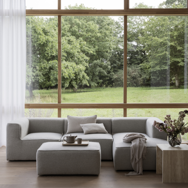 Modernes graues Ecksofa mit gemütlicher Einrichtung und großen Fenstern, die auf eine grüne Landschaft blicken.