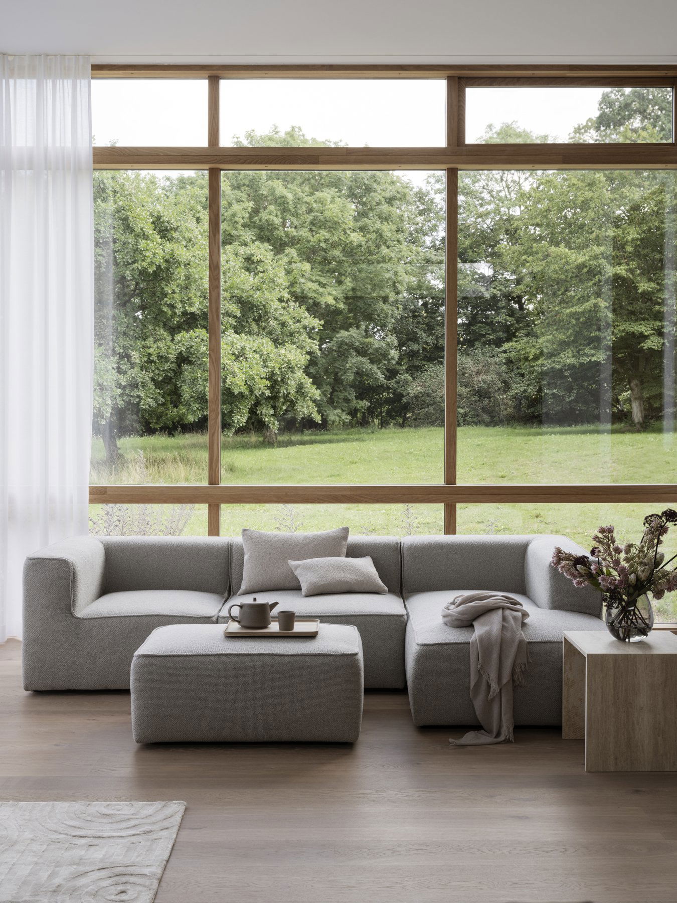 Modernes graues Ecksofa mit gemütlicher Einrichtung und großen Fenstern, die auf eine grüne Landschaft blicken.