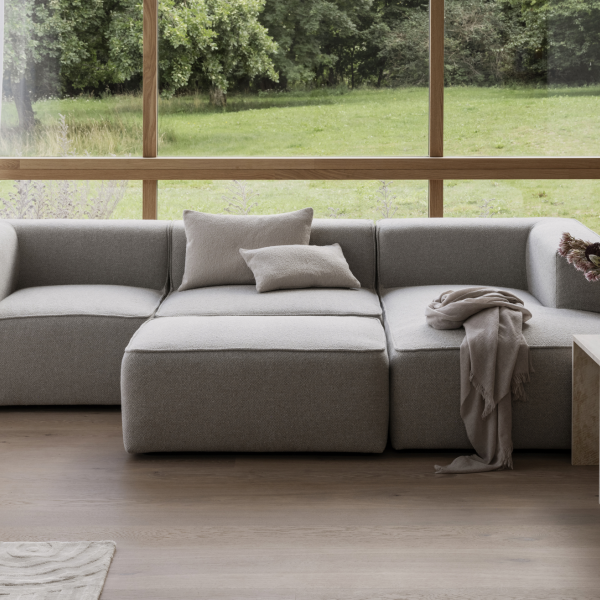 Moderner grauer Ecksofa in einem hellen, luftigen Wohnzimmer mit großen Fenstern und Grünflächen draußen.