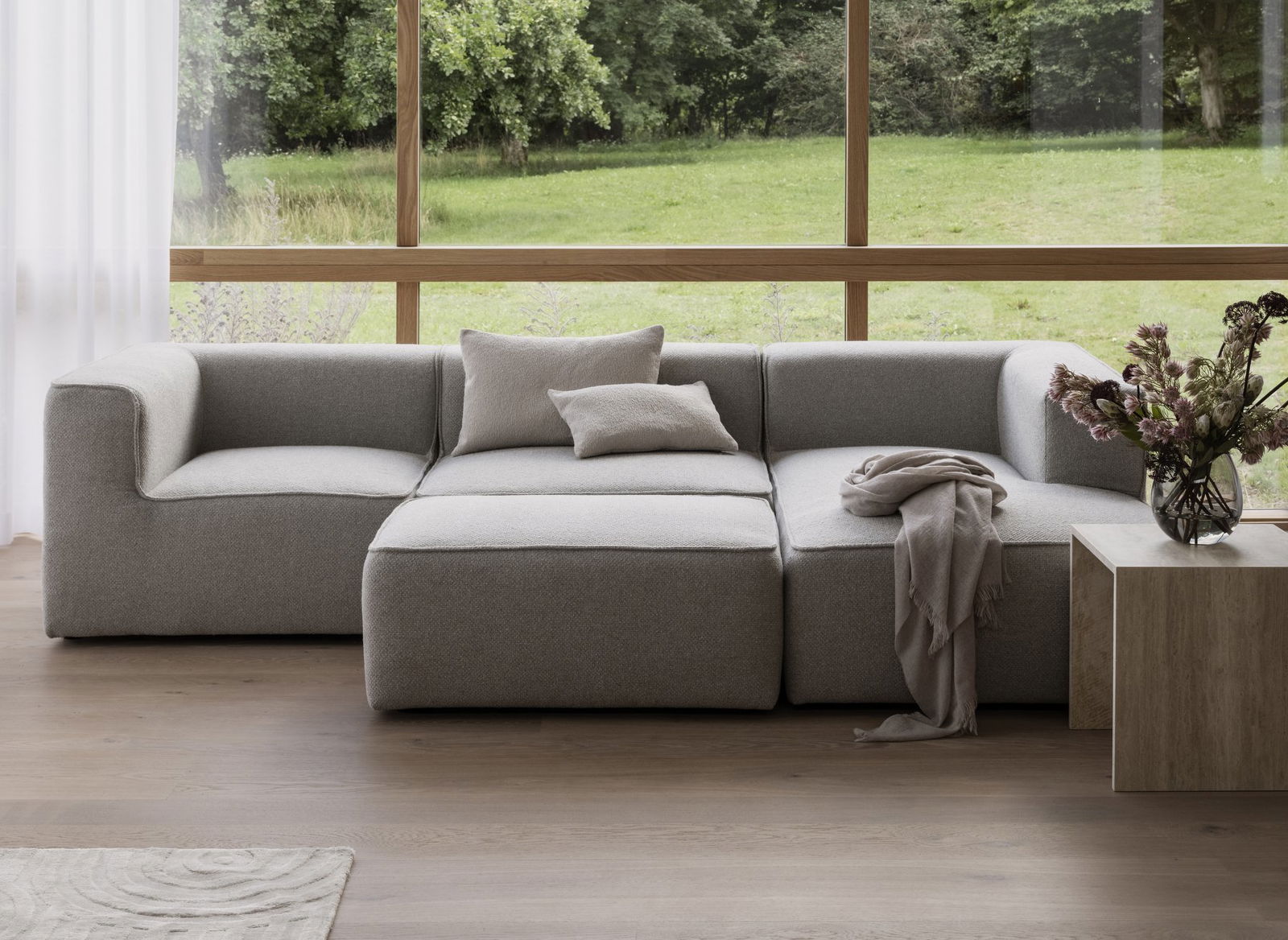Moderner grauer Ecksofa in einem hellen, luftigen Wohnzimmer mit großen Fenstern und Grünflächen draußen.