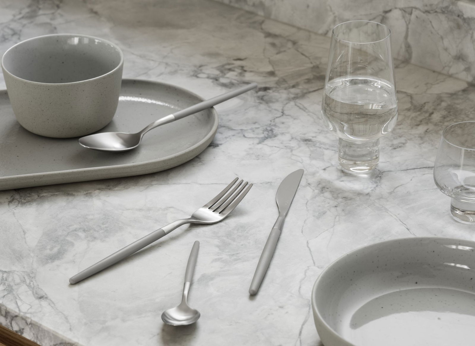 Elegant table setting with modern grey dishware and stainless steel utensils on marble surface.