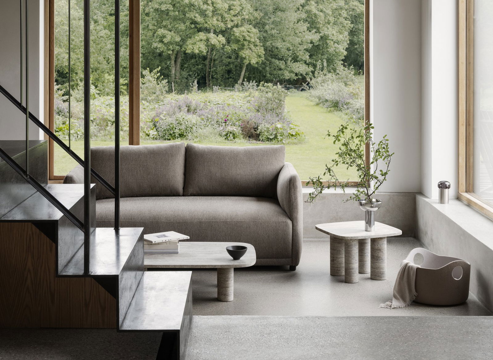 Moderner Wohnraum mit einem grauen Sofa, minimalistischen Möbeln und Blick auf eine üppig grüne Außensicht.