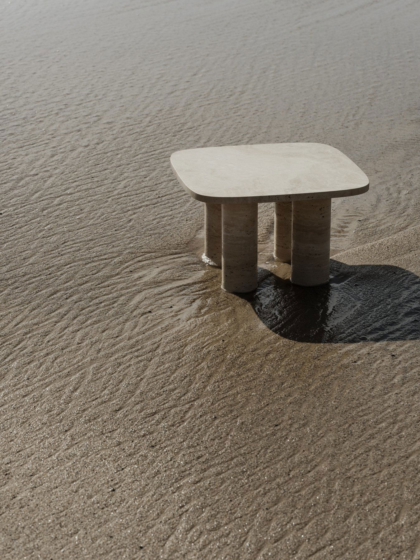 Minimalistischer Steintisch am sandigen Strand mit subtilen Wasserreflexionen.