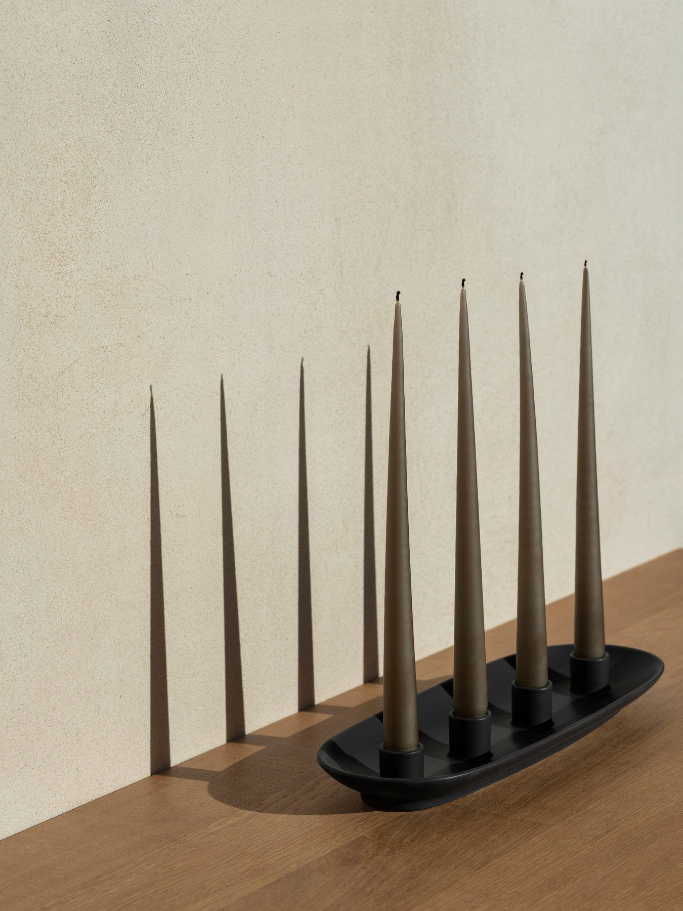 Eleganter Kerzenhalter mit vier konisch geformten Kerzen, die sanfte Schatten an eine strukturierte Wand werfen.