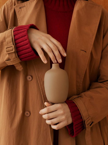 A person holding a minimalist vase while wearing a stylish brown coat and red sweater.