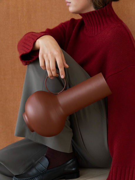 A woman in a red sweater holds a unique brown vase while sitting against a textured backdrop.